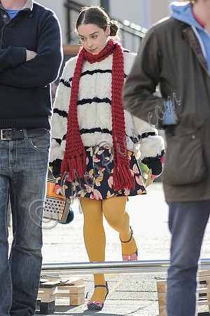 Emilia Clarke on set wearing white with black stripe fur jacket, long red scarf, flowered skirt and yellow tights.