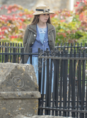 Georgia Brinkworth (Phyll) in the school yard behind railings wearing straw hat
