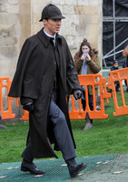 Benedict Cumberbatch wearing long caped coat and deerstalker walking from left to right.