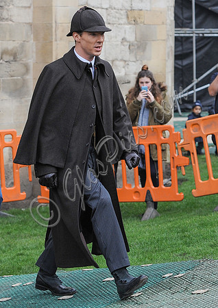 Benedict Cumberbatch wearing long caped coat and deerstalker walking from left to right.