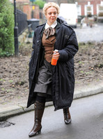 Amanda Abbington carrying orange drink, weraing period costume with long black coat over
