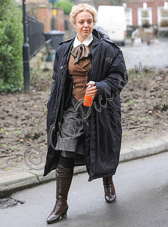 Amanda Abbington carrying orange drink, weraing period costume with long black coat over