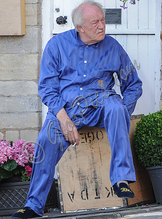 Sir Michael Gambon as Howard Mollison wearing blue pyjamas and monogrammed slippers sat on a tea cheast