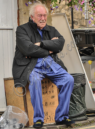 Sir Michael Gambon as Howard Mollison wearing blue pyjamas and monogrammed slippers sat on tea chest