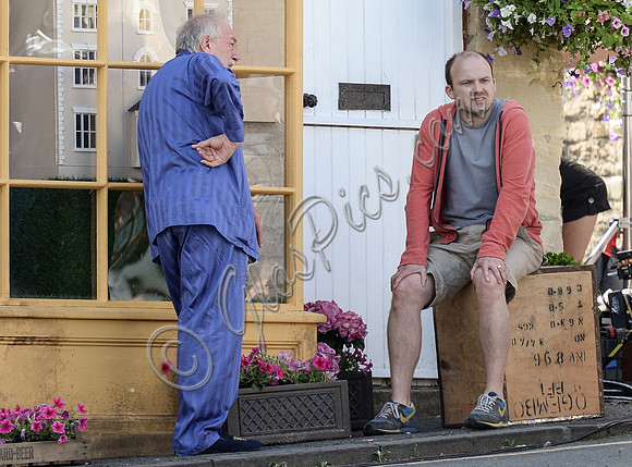 Sir Michael Gambon as Howard Mollison wearing blue pyjamas and monogrammed slippers with Rory Kinnear sat on tea chest