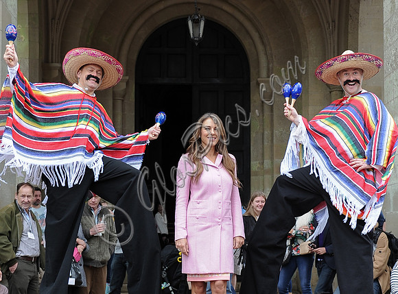 Elizabeth Hurley in pink coat flanked by  two mexicans on stilts wearing ponchos, sombreros and shaking their maracas