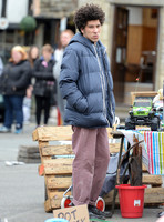 Joel Fry wearing blue padded jacket and pinkish trousers in a market scene during filming "Apocalypse Slough"
