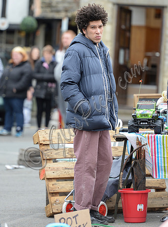 Joel Fry wearing blue padded jacket and pinkish trousers in a market scene during filming "Apocalypse Slough"
