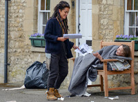Karla Crome with dreadlocks wearing blue jacket, grey trousers and workboots handing missing person notice to rough sleeper on park bench in a scene from "Apocalypse Slough"