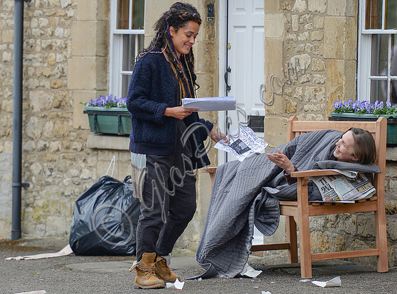Karla Crome with dreadlocks wearing blue jacket, grey trousers and workboots handing missing person notice to rough sleeper on park bench in a scene from "Apocalypse Slough"