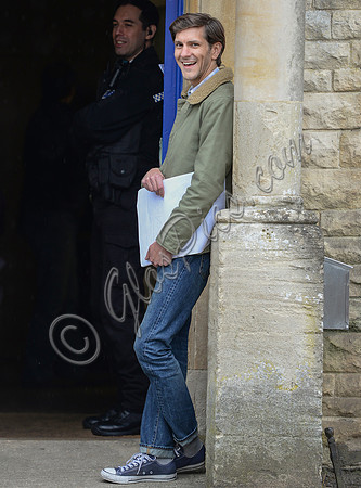 Mathew Baynton leaning against wall, smiling, police officer in background