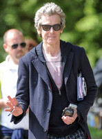 Close up of Peter Capaldi as Doctor Who wearing a long dark jacket and Ray Ban sunglasses waving to camera with pen in hand, and carrying a mobile phone,  and Ray Ban glasses case in other, with pape