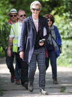Full length shot of Peter Capaldi as Doctor Who wearing a long dark jacket, checked trousers and Ray Ban sunglasses, with others.