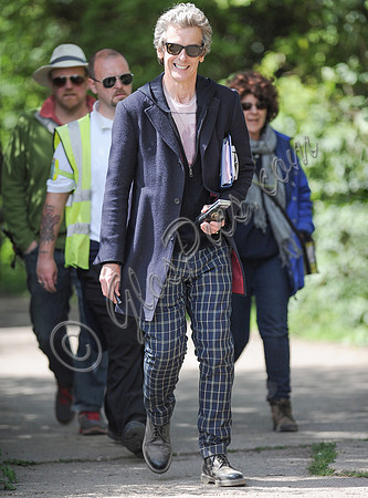 Full length shot of Peter Capaldi as Doctor Who wearing a long dark jacket, checked trousers and Ray Ban sunglasses, with others.