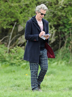 Peter Capaldi as Doctor Who wearing a long dark jacket, checked trousers and Ray Ban sunglasses walking and signing notes.
