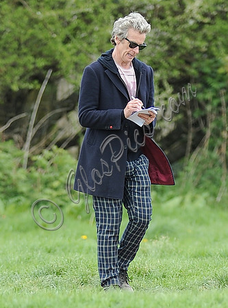 Peter Capaldi as Doctor Who wearing a long dark jacket, checked trousers and Ray Ban sunglasses walking and signing notes.