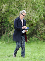 Peter Capaldi as Doctor Who wearing a long dark jacket, checked trousers and Ray Ban sunglasses, carrying a pen, paper and Dr. Who cards