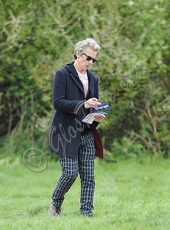 Peter Capaldi as Doctor Who wearing a long dark jacket, checked trousers and Ray Ban sunglasses, carrying a pen, paper and Dr. Who cards