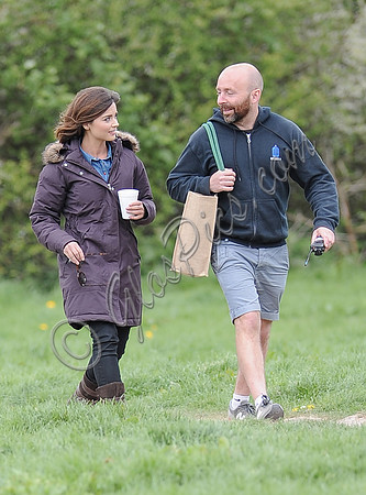Jenna Coleman walking along with crew member carrying her bag,  going on set to film Doctor Who, wearing sunglasses and padded purple coat holding polystyrene cup