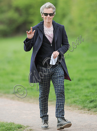 Full length shot of Peter Capaldi as Doctor Who wearing a long dark jacket, checked trousers and Ray Ban sunglasses waving to camera