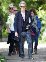 Full length shot of Peter Capaldi as Doctor Who wearing a long dark jacket, checked trousers and Ray Ban sunglasses, with others.