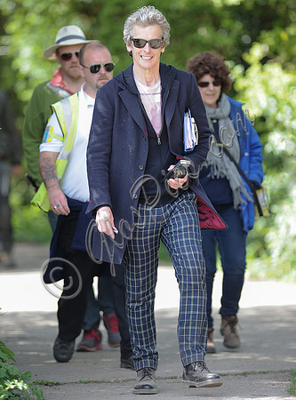 Full length shot of Peter Capaldi as Doctor Who wearing a long dark jacket, checked trousers and Ray Ban sunglasses, with others.