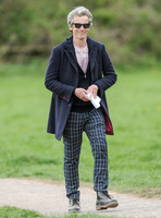 Full length shot of Peter Capaldi as Doctor Who wearing a long dark jacket, checked trousers and Ray Ban sunglasses