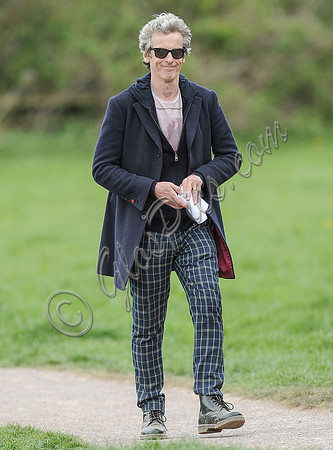 Full length shot of Peter Capaldi as Doctor Who wearing a long dark jacket, checked trousers and Ray Ban sunglasses