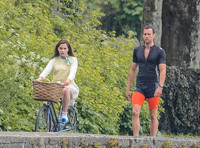 Emilia Clarke riding bicycle wearing lemon floral dress, lemon jumper and turquoise neckerchief alongside Matthew Lewis in orange and black running gear