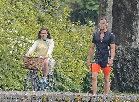 Emilia Clarke riding bicycle wearing lemon floral dress, lemon jumper and turquoise neckerchief alongside Matthew Lewis in orange and black running gear