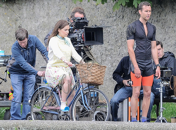 Emilia Clarke riding bicycle wearing lemon floral dress, lemon jumper and turquoise neckerchief alongside Matthew Lewis in orange and black running gear