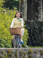 Emilia Clarke on bicycle with handlebar basket wearing lemon floral dress, lemon jumper and turquoise neckerchief