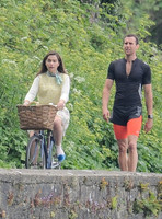 Emilia Clarke riding bicycle wearing lemon floral dress, lemon jumper and turquoise neckerchief alongside Matthew Lewis in orange and black running gear