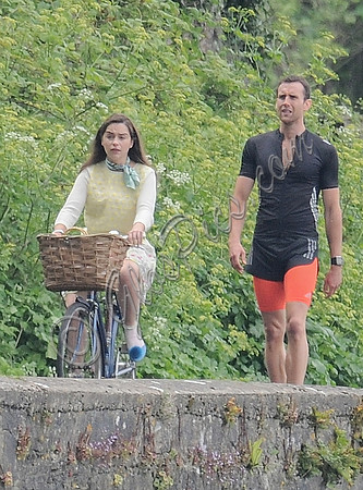 Emilia Clarke riding bicycle wearing lemon floral dress, lemon jumper and turquoise neckerchief alongside Matthew Lewis in orange and black running gear