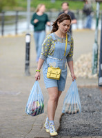Emilia Clarke on set carrying two plastic varrier bags of shopping, wearing pale blue dungaree dress with heart motif, pineapple print jumper, blue ankle socks with lemonT bar shoes and a yellow mini