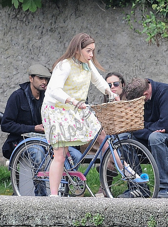 Emilia Clarke looking shakey on bicycle with handlebar basket wearing lemon floral dress, lemon jumper and turquoise neckerchief