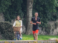 Emilia Clarke riding bicycle wearing lemon floral dress, lemon jumper and turquoise neckerchief alongside Matthew Lewis in orange and black running gear
