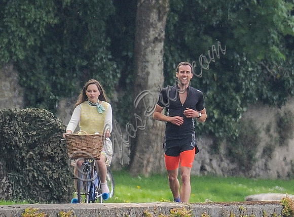 Emilia Clarke riding bicycle wearing lemon floral dress, lemon jumper and turquoise neckerchief alongside Matthew Lewis in orange and black running gear