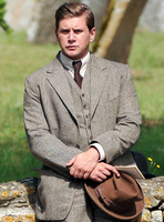 Allen Leech in tweed suit carring hat sat on cotswold stone wall