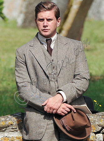 Allen Leech in tweed suit carring hat sat on cotswold stone wall