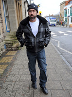 A bearded Nicolas Cage wearing plaid jacket and beanie hat, walking alone in Glastonbury, Somerset.