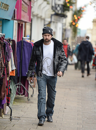 A bearded Nicolas Cage wearing plaid jacket and beanie hat, walking alone in Glastonbury, Somerset.