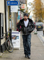 A bearded Nicolas Cage wearing plaid jacket and beanie hat, passing under blue library sign in Glastonbury, Somerset,