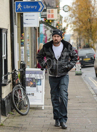 A bearded Nicolas Cage wearing plaid jacket and beanie hat, passing under blue library sign in Glastonbury, Somerset,