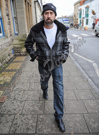 A bearded Nicolas Cage wearing plaid jacket and beanie hat, walking alone in Glastonbury, Somerset.