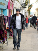 A bearded Nicolas Cage wearing plaid jacket and beanie hat, walking alone in busy Glastonbury High Street.