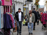A bearded Nicolas Cage wearing plaid jacket and beanie hat, walking and talking to leather artisan in flat cap in  Glastonbury High Street, amongst others.
