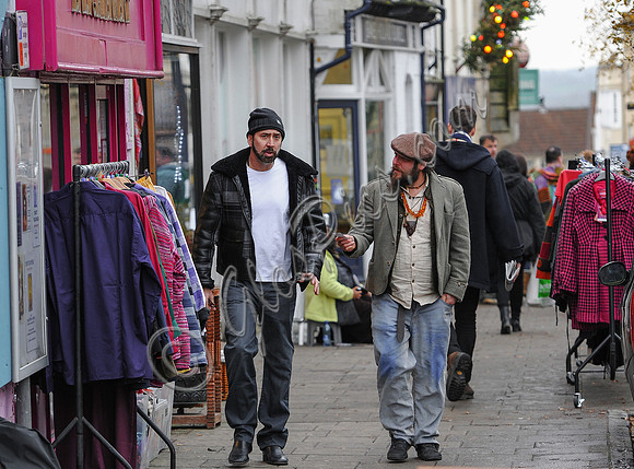 A bearded Nicolas Cage wearing plaid jacket and beanie hat, walking and talking to leather artisan in flat cap in  Glastonbury High Street, amongst others.