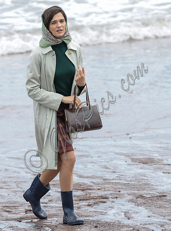 Rachel Weisz on beach carrying handbag, wearing 60's style pale green raincoat, headscarf and blue short wellington boots