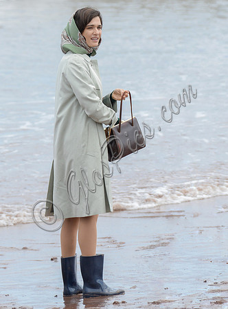 Rachel Weisz on beach carrying handbag, wearing 60's style pale green raincoat, headscarf and blue short wellington boots